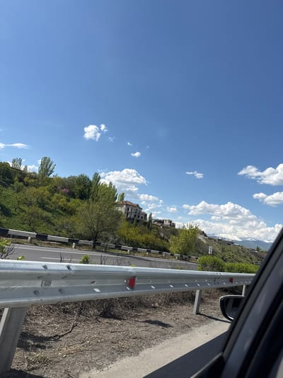 The image, taken from within a vehicle, shows a roadside scene in Getamej, Armenia on a clear day with a blue sky dotted with scattered white clouds. A metal guardrail runs parallel to the asphalt road, with a grassy slope dotted with trees beyond it. Several buildings are visible in the distance. The car's side mirror and window frame are partially visible in the frame.