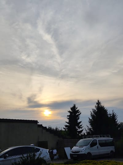 The image shows a residential area in Czapury, Poland, with a view of the sky during sunset or sunrise. In the foreground, there is a white car, possibly a taxi, parked on the left side, partially obscured by green foliage. To the right, a white van is parked. Behind the vehicles, a wall runs horizontally across the frame, with a building or structure behind it. Several tall evergreen trees are silhouetted against the sky. The sky itself is filled with clouds, with the sun visible as a bright, diffused light source low on the horizon, casting a warm glow. There are no people visible in the image. The environment appears to be a semi-rural or suburban setting.