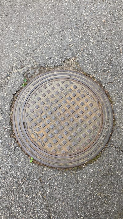 An overhead shot shows a circular, rusty metal manhole cover embedded in cracked asphalt. The manhole cover features a grid pattern of raised squares and has text inscribed around its circumference. On the left side, it reads "uponor" in a curved font, followed by "EN®124 D400" in a straighter font. On the right side, the text "uponor" is also visible in a curved font, and further around, it reads "EN®124-D400 L". Small green weeds are growing in the cracks of the asphalt surrounding the manhole cover. The environment is an outdoor urban setting, likely a street or sidewalk in the city of Ūlupji, Latvia. There are no people or animals visible in the image.