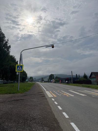 The image shows a street scene in Krasnyy Klyuch, Russia, under an overcast sky with the sun peeking through the clouds. On the left side of the road, a pedestrian crossing sign with a yellow triangle and a blue square depicting a pedestrian is mounted on a pole. A traffic light with a single orange signal is suspended over the road. The road itself has a pedestrian crossing marked with white and yellow stripes. In the background, there are houses and buildings lining the street, with trees and a misty mountain range in the distance. The road is asphalt and appears to be in a rural or semi-rural area. No people are visible in the image.
