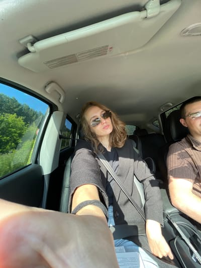 A woman wearing sunglasses and a black jacket is seated in the passenger seat of a car, holding up her left arm, likely to take a selfie. She is wearing a seatbelt. Next to her, a man wearing glasses and a brown shirt is in the driver's seat. The car's interior is visible, including the sun visor. Through the window, a treed landscape with a bright blue sky can be seen. The setting is likely within or near the city of Shud'ya, Russia, given the context of the image's origin.