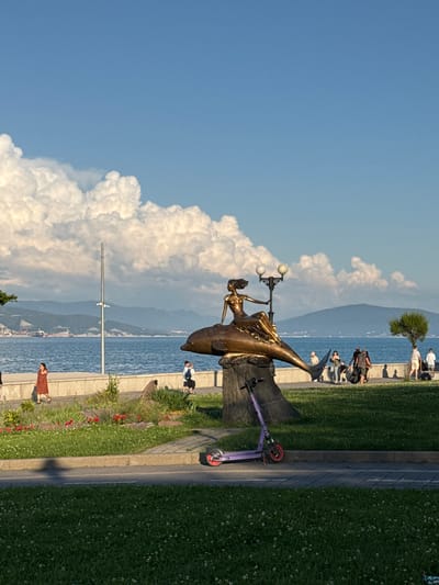 A bronze sculpture of a woman with flowing hair sitting on a dolphin is prominently displayed in a park setting. The sculpture is positioned on a stone base with a lamppost behind it. To the left of the sculpture, a woman in a reddish-brown dress walks along a paved pathway. Further down the pathway, a group of people, including children, are visible, some walking and others standing. A purple electric scooter is parked on the grass to the left of the pathway. In the background, a body of water, likely the sea, stretches towards distant mountains. The sky above is a clear blue with large, fluffy white clouds. The park area features green grass, some scattered flowers, and a few trees. The overall scene depicts a public space by the water, with people enjoying a leisurely day outdoors.