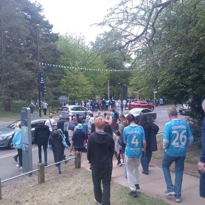 The image captures a scene in Coventry, United Kingdom, specifically at the War Memorial Park near Top Green Park. A sizable crowd of people has gathered along a road. Banners with blue and white triangular flags are strung across the road, creating an atmosphere of celebration or event.
Many individuals are wearing light blue shirts, with two prominently displaying the names "THOMAS" with the number "4" and "CHAMPIONS" with the number "26," along with the logo "monzo". These suggest the crowd might be associated with a sports team or organization.
A sign with the text "CCFC CHAMPIONS #FLISB" indicates the celebration is possibly related to the Coventry City Football Club (CCFC) winning a championship.
Cars are visible on the road, including silver, red, and black vehicles. On the side of the road is a sign that says "WE ARE BACK". The backdrop consists of trees and greenery typical of a park environment.