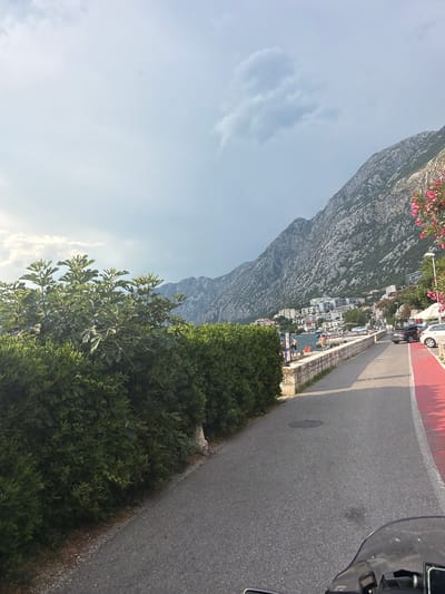 A road runs alongside a stone-walled embankment, leading into a coastal town nestled at the base of a mountain range. The road has a red-painted lane on the right side. Lush green bushes line the left side of the road, with trees further back. Across the embankment and water, buildings are visible on the mountainside, suggesting a town or village. People are seen on the embankment near the water and on the beach. Several cars are parked along the road. The sky is overcast with scattered clouds. The scene appears to be in Dobrota, Montenegro, given the distinctive mountainous coastal landscape.