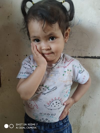 A young child poses for a photo. The child has dark hair styled in two pigtails with yellow hair ties. They are wearing a patterned shirt with dinosaurs and the words "Velvet Junior" on it. They are also wearing blue jeans. The child has one hand on their cheek and the other on their hip. The background appears to be a wall with some markings. The bottom of the image includes a camera watermark.