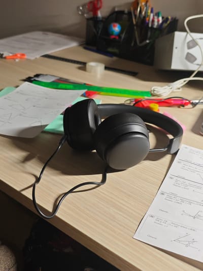 This media file shows a close-up, eye-level view of a wooden desk with various school supplies. The prominent objects include a pair of black headphones with a coiled black wire, lying on top of several sheets of paper. One sheet of paper, in the foreground, contains geometry problems with diagrams. Other items on the desk include rulers (one green, one red), pens, pencils in a holder, a small globe, scissors, and a roll of clear tape. In the background, a white electronic device with a coiled white wire is visible. The lighting suggests it might be nighttime or a dimly lit room, as there are strong shadows cast by the headphones and wire. The scene depicts a study or work area, possibly at home, with materials laid out for learning or homework. The visible text is in Russian and pertains to geometry problems, with labels such as "Дано" (Given), "Найти" (Find), and specific angles and line segments denoted. The overall mood is one of focused study or a moment of pause during an academic task. No people are visible. The location context of Svietlahorsk, Belarus is not visually evident in the image itself.