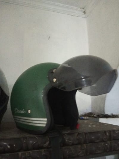 The image depicts an indoor scene in B. Nam Ngao, Laos. The primary subject is a green, open-face motorcycle helmet positioned centrally, featuring a raised, dark-tinted visor. The helmet's left side displays the word "Classic" in white lettering and three horizontal white stripes. To the immediate left, a portion of a second, dark-colored helmet is partially visible. Both helmets are situated on a dark surface, possibly wood, which exhibits carved or embossed patterns. On this surface, to the right of the green helmet, a small red object, a light-colored cylindrical item, and a cluster of small metallic objects are present. The background consists of a plain white wall with a white crown molding running along the top edge. No individuals are depicted, and no active interactions are occurring.