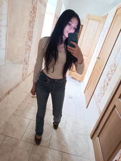 A woman stands in a tiled bathroom, holding a smartphone to take a selfie. She is wearing a light-colored long-sleeved top, blue jeans, and brown shoes. The bathroom has light-colored tiles with decorative borders. There are wooden doors visible. The exact location in Simferopol, Ukraine, cannot be determined with certainty from the image alone.