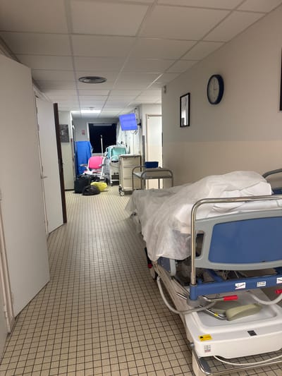 The image depicts the interior of a hospital hallway, likely in Tourcoing, France given the instructions, though no direct markers of the city are visible. The hallway is lined with white tiled walls on the left and patterned tile walls on the right. Several hospital beds, covered with disposable sheets and possibly awaiting cleaning or transport, are lined up along the right side. One bed closest to the camera has a white sheet draped over it. Further down the hall, there is a blue topped bed and trash bags. On the left wall, there's a wall clock. A closed white door is in the foreground. No people are visible in the frame.