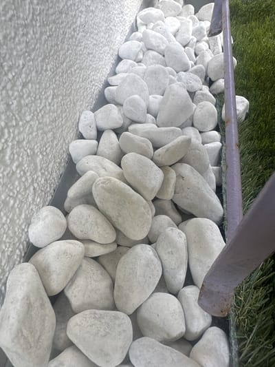 The image shows a close-up, low-angle view of a collection of smooth, white river stones. These stones are densely packed and form a textured surface along the bottom edge of the frame. To the left, a coarse, light-colored granular surface, possibly gravel or rough concrete, fills the lower portion of the image.

Behind the stones, a thin, purple metal bar runs horizontally across the middle of the frame, appearing to be part of a railing or support structure. Beyond the bar, a patch of artificial green turf is visible, suggesting an outdoor setting like a garden or a recreational area. The lighting suggests it is daytime, and there are no indicators of specific weather conditions.

The scene is devoid of people or any discernible activity. The focus is entirely on the arrangement of natural and man-made elements, highlighting their textures and forms. The stones are varied in size and shape, some rounded and others more elongated, creating a visually engaging pattern. The overall impression is one of neatness and perhaps a decorative landscaping element. The location context of Hamburg, Germany, provides a geographical reference but does not offer specific visual cues within the image itself. No text is visible in the image.