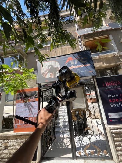 The image, captured from a low-angle perspective, depicts the entrance of a building complex in As-Salihiya Municipality, Syria, under bright daylight. A human arm is visible in the lower-middle portion, holding a black camera gimbal with "RS4" visible on its structure. A yellow camera body with a black lens is mounted on the gimbal, pointing upwards towards the building's facade and signs.
Dominating the upper-middle is a large, dark rectangular sign for 'معهد النهضة الأكاديمي' (Al-Nahda Academic Institute), featuring white and orange 'ATC' lettering. On the left side of this sign, an illustration shows a person in traditional attire, possibly a keffiyeh, holding what appears to be a rifle or shotgun, with another individual partially visible behind them. Below this, smaller Arabic text is visible. To the left of the main entrance, a tall, upright poster titled 'دورة صيف' (Summer Course) advertises academic programs for 2026-2027, displaying an image of multiple hands holding diplomas. Arabic text details specific subjects and a QR code is present at the bottom. To the right, another dark banner displays 'LINGUA Learn to speak English,' indicating an English language center affiliated with the University of Cambridge and ETS Global, with an Instagram handle visible.
The building itself is multi-storied with light-colored exterior walls. Multiple windows with external roller shutters are visible on various levels, and an LG air conditioning unit is affixed to a wall. An upper balcony features two dark-colored solar panels. A decorative black wrought iron gate with gold-painted accents, adorned with two empty picture frames, blocks the entrance, behind which light-colored stairs ascend. Lush green foliage with long, narrow leaves hangs overhead, partially obscuring the upper portion of the building and casting dappled shadows across the scene. A clear blue sky is visible through the gaps in the leaves.