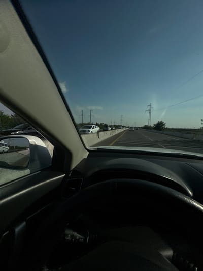 The image is taken from the perspective of someone driving a car on a highway in Istiqlol, Uzbekistan. The highway is congested with traffic, with numerous cars visible on the road. A concrete barrier separates the lanes of traffic. In the distance, power lines and transmission towers are visible against a clear blue sky with a few wispy clouds. The interior of the car is partially visible, including the steering wheel and part of the dashboard. The side mirror reflects a white van driving in the opposite direction. The sun appears to be high in the sky, suggesting it is daytime.