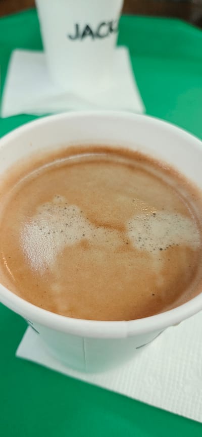A close-up, eye-level shot shows the surface of a coffee drink in a white paper cup. The drink has a light brown, foamy crema with small bubbles. The cup is placed on a white paper napkin, which rests on a bright green surface, likely a table. In the blurred background, another white paper cup with "JACK" printed in black is visible. The scene appears to be indoors.
