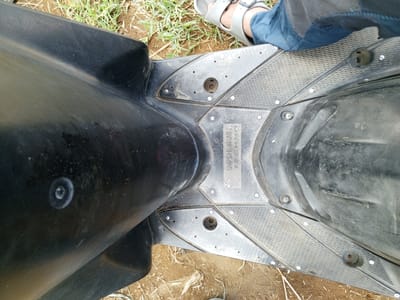 A top-down shot displays the footrest of a black scooter on a patchy ground surface. A person is visible to the upper right, wearing blue pants and gray sandals. The footrest is grimy and textured, secured with numerous screws. A rectangular plate labeled "YOSHIMURA TECHNO" is positioned in the center. The background includes sparse green foliage and bare soil. There is no clear evidence to pinpoint the exact location in Ban Katan, Laos.