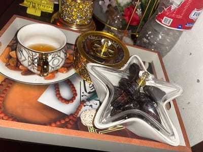 A tray holds a star-shaped container filled with dates, a golden ornate jar, and a patterned teacup with almonds on a saucer. The tray has a decorative background featuring prayer beads. In the foreground, there's a red container, a plastic bottle, gold stands, a jeweled candle holder, and yellow hair clips. The scene appears cluttered and is captured from a high angle.
