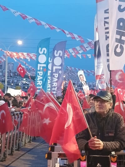 The image shows a gathering of people in a public space, most likely a political rally or demonstration. The event is held in Istanbul, Turkey at the coordinates 41.084 latitude and 28.678 longitude. The image depicts a large group of people holding flags. Several people are visible in the foreground, many wearing red and white colors. Several flags are hanging across the image, including Turkish flags and banners with the text "SOL PARTI" and "CHP." The foreground subject is a middle-aged man wearing a black jacket and glasses, holding a Turkish flag. He appears to be talking with someone to the right of the frame. In the background, there are numerous other people, most of them holding red and white flags and banners, and a few with the CHP flags. The environment is likely a city park or a public square. The scene is dominated by the flags and the crowd, with the cityscape in the background. The lighting is relatively low, suggesting that the image was taken in the late afternoon or early evening.