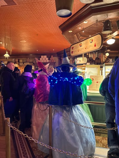 The image shows a whimsical display inside a shop, likely related to Harry Potter, as evidenced by the wizard hats. The scene features colorful, layered wizard costumes arranged on what appears to be a suspended display rack with chains. The hats are adorned with strings of lights. People are looking at the display. There's a menu board on the ceiling and shelves full of merchandise in the background. The setting gives off a fantastical and enchanting atmosphere.