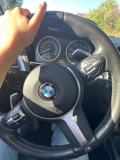 The image shows a close-up of a person's hands on the steering wheel of a car, as seen from the driver's perspective. The steering wheel is black leather with silver accents and features the BMW logo in the center. On the left side of the steering wheel, there are control buttons for volume and mode, and on the right, there are buttons for cruise control and limiter functions. The car's dashboard instruments, including a speedometer, are visible behind the steering wheel, with the needle on the speedometer indicating a speed of approximately 120 km/h. The sunlight suggests it is daytime, and the clear sky implies good weather. The background is blurred, but hints of green trees and a bright sky suggest an outdoor setting, likely a road. The overall impression is of someone driving on a sunny day. Visible text includes "BMW" on the steering wheel and "M" logo on the steering wheel. There is also some text on the dashboard instruments which is not fully legible. The scene is set in Gorni Bogrov, Bulgaria.