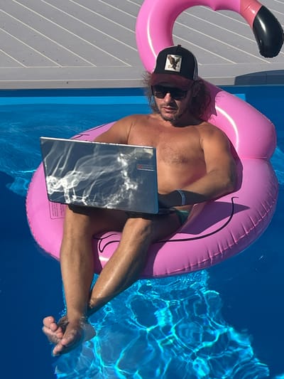 A shirtless man wearing a baseball cap and sunglasses is sitting on a pink inflatable flamingo in a swimming pool. He is holding a silver laptop on his lap and appears to be working. The swimming pool is surrounded by a wooden deck. The man has long, curly brown hair and is wearing swim trunks. The water in the pool is blue and clear, with sunlight reflecting on the surface. The man has a bracelet on his left wrist. The scene takes place outdoors on a sunny day.