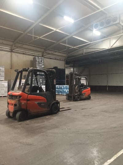 The image depicts the interior of a warehouse in Sexbierum, Netherlands. The space is industrial, with a concrete floor and a high ceiling supported by metal beams. Fluorescent lights illuminate the scene, along with a large air conditioning unit mounted on the ceiling at the far end.
In the foreground, an orange and black Linde forklift is parked, its tines extended. It appears to be dirty, suggesting recent use. In the mid-ground, another forklift, also orange and black, is positioned further into the warehouse.
The warehouse is stocked with goods. On the left, stacks of palletized items wrapped in plastic are visible. In the center and right, rows of black plastic crates and palletized boxes, likely containing bottled water, are neatly arranged. The overall impression is one of a functional, operational storage facility.