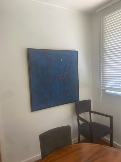 The image shows a dining room scene from a slightly elevated angle, oriented vertically. A dark-framed, abstract painting with a deep blue background and scattered red and white elements hangs on a white wall. To the right of the painting, a modern wooden dining table with a rich grain is partially visible, along with two dark gray chairs with wooden frames. The chairs have curved seats and backrests. One chair is positioned next to the table, and another is further to the right, facing away from the camera. Above the painting and to the left, white horizontal blinds cover a window, suggesting it is daytime. The overall impression is one of a contemporary and minimalist interior. There is no visible text in the image. The location context is Matosinhos, Portugal.