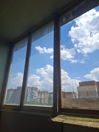 The image is taken from an interior perspective looking out through a window, likely at an apartment building in Donetsk, Ukraine. The view is divided into two sections by a horizontal wooden beam. The top section shows a bright blue sky filled with fluffy white clouds, suggesting it is daytime and the weather is fair. A fine mesh screen is visible in front of the sky in this upper section. On the right side of the upper section, the facade of a brick apartment building is visible.
The lower section of the window also looks out onto an apartment building, but this view appears slightly more distant and is framed by the window structure itself. Reflections on the glass might be present, but the primary view is of the building. The wooden window frame appears old and somewhat worn, with peeling paint in some areas. The overall scene evokes a sense of everyday life in an urban environment, with a clear and sunny sky overhead. There is no visible text in the image.