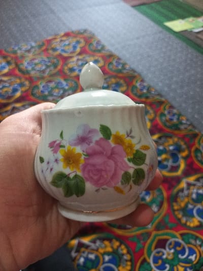 The image displays a small, white, lidded ceramic container held in a human hand, located in Navbahor, Uzbekistan. The container features a rounded body, a short pedestal base, and a domed lid with a pointed finial. It is decorated with a floral motif comprising pink roses, yellow blossoms, purple accent flowers, and green leaves, with a thin gold band at its base. A light-skinned human hand grips the container from the left side. The background consists of a red textile with an intricate repeating pattern in yellow, blue, and green. Behind this, a grey, textured surface is visible, with a green object and light-colored papers partially discernible in the upper right background.