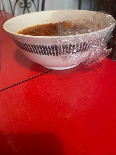 The scene is located in Fegersheim, France. A white ceramic bowl, featuring a continuous band of dark, stylized leaf patterns around its exterior, occupies the upper portion of the frame. The bowl contains a reddish-brown, semi-liquid food substance and is partially covered by clear, wrinkled plastic wrap. It rests on a vivid red, textured surface that exhibits a linear seam extending horizontally across the lower-left area. The background is out of focus, revealing indistinct shapes and a dark, decorative metal scrollwork element in the upper section. No individuals, explicit actions, or direct interactions are depicted within the image.