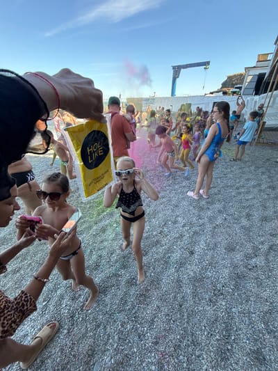 This media file captures a lively scene at what appears to be a beach festival, possibly a color powder event, given the presence of colorful powders in the air and on people. The subjects are a diverse group of people, including children and adults, dressed in swimwear and casual clothing. Some individuals are holding phones, suggesting they are documenting the event. In the foreground, a hand is holding a yellow bag with black text that reads "HOLI LIKE" and some Cyrillic text.
The setting is a pebble beach, with a blue sky overhead indicating it is daytime, likely sunny. In the background, there are some structures that could be part of a beach resort or entertainment venue, including what looks like a crane. The atmosphere appears joyful and energetic, with people actively participating in the event. The presence of colorful powders, some of which are visible as pink clouds, strongly suggests a Holi-like celebration where participants throw colored powder at each other. The overall mood is festive and celebratory.