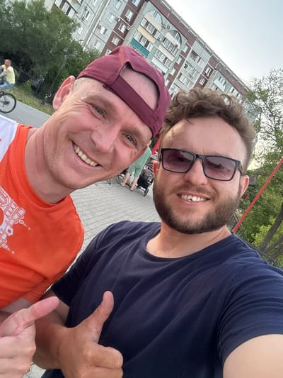 This is a selfie taken by two adult males outdoors, possibly on a pedestrian walkway or promenade, with an apartment building in the background. The man on the left is wearing a maroon baseball cap and an orange t-shirt with white trim. He is smiling and has a slight tan. The man on the right is wearing sunglasses and a dark blue t-shirt. He has a beard and is also smiling. Both men are giving a thumbs-up gesture. In the background, a person on a bicycle is visible in the distance, and further back, a multi-story apartment building with arched windows can be seen. Trees and a red railing are also part of the environment. The pavement appears to be made of interlocking concrete pavers. The overall impression is a casual, outdoor scene, likely in a populated urban area like Blagoveshchensk, Russia, given the architectural style of the building and the presence of greenery and pedestrian infrastructure.