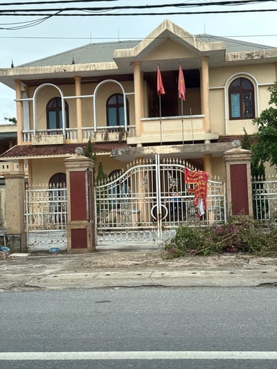 The image shows a large, light yellow, two-story house with a dark gray, corrugated metal roof. The house has a covered balcony on the second floor, supported by beige columns. Two red flags with yellow stars are flying from poles on the balcony. The first floor has an arched entryway with a white metal gate. The gate is decorated with intricate scrollwork and floral motifs. A red banner with yellow writing is hanging from the gate. In front of the gate, there is a pile of green branches with pink flowers. To the left of the gate, there is a beige stone pillar and to the right, a similar pillar with red accents. The house is situated on a street with a gray asphalt road. The sky is overcast and appears to be a light gray. The location is specified as Triệu Phong Commune, Vietnam.