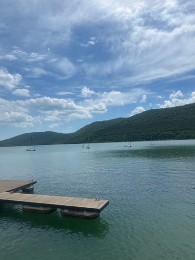 The image depicts a scenic view of a body of water, likely Abrau Lake, in Abrau-Dyurso, Russia. In the foreground, a wooden pier extends into the tranquil blue water. The pier is constructed with wooden planks and supported by buoys. Several sailboats are visible on the lake, with their masts piercing the sky. In the background, lush green hills covered in trees rise majestically, forming a natural backdrop to the serene landscape. The sky above is a brilliant blue, adorned with fluffy white clouds, suggesting a clear and pleasant day.