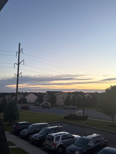 The image captures a twilight scene in Allen Township, United States of America. A clear sky with a gradient of blue to pale yellow dominates the upper portion, with wispy clouds tinged with orange and pink along the horizon. Prominent utility poles with power lines stretch across the frame. In the mid-ground, a residential neighborhood features a line of houses with visible roofs and windows, nestled amongst trees. A street runs through the neighborhood with several parked vehicles, including a red pickup truck in motion. In the foreground, a parking lot is filled with several parked cars, including black SUVs and a silver SUV. A signpost for "MR" and "LEASING CENTER NOW OPEN" is visible near the street. The overall impression is of a quiet suburban evening.
