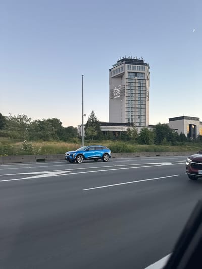 The image shows a view from a moving vehicle on a highway in Burnaby, Canada, looking towards the Grand Villa Casino. In the foreground, a blue SUV is driving in the left lane, while a red vehicle is partially visible in the right lane. Beyond the highway, a tall, modern building with "DELTA" and "Grand Villa casino" branding is prominent. The sky is clear and light blue with a crescent moon visible. Green trees and vegetation line the highway. The scene appears to be captured during the late afternoon or early evening.
