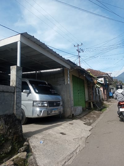 A daytime street scene captures a semi-urban environment under a clear blue sky with scattered clouds. In the foreground-left, a silver Suzuki Carry van, displaying an Indonesian license plate (B 9709 TR), is parked beneath a corrugated metal carport supported by concrete pillars and an unpainted cinder block wall. A cracked concrete apron with a discarded plastic bottle provides access from the street to the carport. Adjacent to the carport, to the right, is a building with closed green louvered doors, followed by a series of older, single-story structures, some with visible deterioration. A prominent utility pole laden with numerous overhead wires stands in the mid-ground, while a distant mountain range is visible against the horizon. On the right side of the asphalt road, a single individual wearing a grey long-sleeved garment and a black helmet is seen from behind, operating a dark-colored motorcycle and moving away from the camera. The requested location is Phonengam, Laos; however, the vehicle's license plate points to an Indonesian origin.