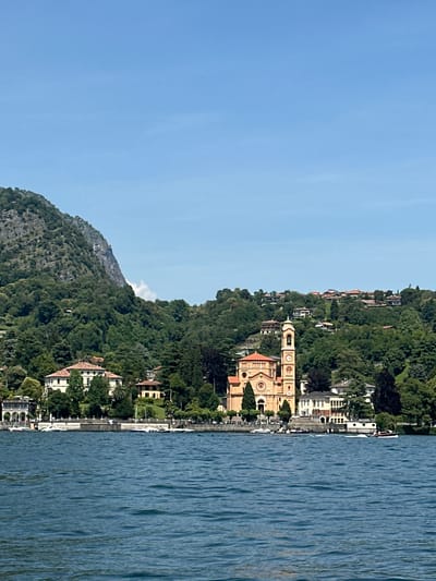 This is a full-frame, eye-level, outdoor, horizontal photograph taken from a body of water, likely Lake Como in Tremezzina, Italy. The scene depicts a shoreline dotted with buildings and lush green trees. In the center-left of the frame, a prominent bell tower and a Romanesque-style church with a red tiled roof stand out. The church is painted a light orange-yellow and has circular windows. To the left of the church, a large white villa with arched windows and a terracotta roof is visible, partially obscured by trees. Further left, other residential buildings with terracotta roofs are nestled along the shore. A steep, rocky mountain covered in dense green trees dominates the background on the left side of the image, with a lighter rocky face visible near its peak. The right side of the background shows a hillside covered with more trees and scattered houses with red roofs. The foreground is filled with deep blue, choppy water, suggesting a breezy day. A white motorboat with two visible occupants is cutting through the water on the right side, creating a wake. Several other smaller boats are docked or moving slowly along the shoreline. The sky is a clear, light blue with a few wispy white clouds on the horizon.