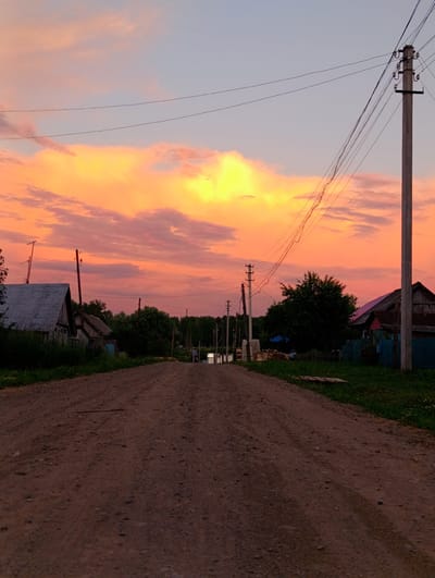 The image captures a rural landscape in the village of Zipunovo, Russia, during a vibrant sunset. A dirt road stretches into the distance, flanked by sparse vegetation and several rustic wooden houses. Power lines traverse the scene, supported by utility poles, with one prominent pole in the foreground on the right. The sky is a dramatic spectacle of orange, pink, and yellow clouds, illuminated by the setting sun, against a soft blue backdrop. A lone figure can be seen in the distance, walking along the road. The overall impression is one of quiet rural life against a breathtaking natural backdrop.