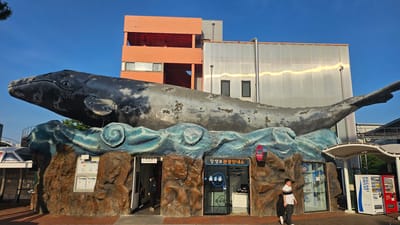 The image shows the Jangsaengpo Whale Culture Village in Ulsan, South Korea. A large, metallic whale sculpture sits atop a building designed to resemble rocky waves, creating the appearance of a whale emerging from the sea. The base of the building incorporates several doorways, one marked "RESTROOM" with a restroom icon, and another with signage in Korean, identifiable as the visitor information center. A man is standing in front of one of the entrances wearing a white and navy-striped shirt. There is also a person entering the restroom. To the right of the main structure, there's a white structure with a vending machine. The building behind the whale sculpture is a mix of orange and silver structures. The sky is clear blue.