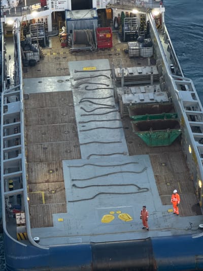 This is an aerial, high-angle, medium shot photograph taken on the deck of a large, industrial vessel at sea. The vessel appears to be a supply or cargo ship, with a deck covered in wooden planks and metal grating. There are several large, green skip bins on the deck, filled with debris. A coiled rope is also visible on the deck, as well as some yellow markings. Two individuals, wearing orange safety suits, are standing on the deck. One is facing away from the camera, while the other is facing the camera. The water surrounding the ship is a dark blue. The lighting suggests it is daytime, but the sky is not visible. The scene is static and appears to be an operational environment on a commercial vessel. There is no indication of the location being in a city or in Italy.