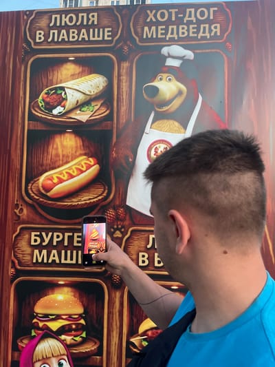 The image shows a person holding a smartphone and taking a picture of a large, wooden-themed food advertisement. The advertisement features cartoon images of food items and a cartoon bear wearing a chef's hat. The text on the advertisement is in Russian and appears to be related to fast food. Specifically, the visible text includes "ХОТ-ДОГ МЕДВЕДЯ" (Bear's Hot Dog), "ЛЮЛЯ В ЛАВАШЕ" (Lyulya in Lavash), and "БУРГЕР МАШИ" (Burger Masha). The smartphone screen also displays an image with similar Russian text, suggesting it might be a game or another advertisement.

The setting appears to be outdoors, possibly in Shcherbinka, Russia, given the context provided. The lighting suggests it is daytime. The person is wearing a blue t-shirt and is angled towards the advertisement, engrossed in taking the photo. The overall scene conveys a casual, everyday moment of someone capturing an image of an interesting advertisement. The advertisement itself is vibrant and designed to be eye-catching, with warm, earthy tones dominating the display. The cartoon characters and food imagery create a playful and inviting atmosphere.