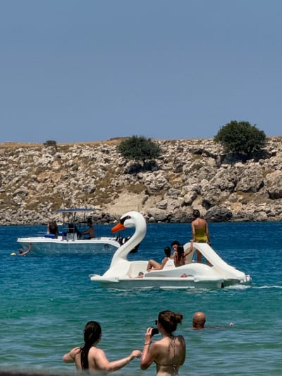 This is a medium shot of a bright, sunny day at a beach in Lindos, Greece. The water is a vibrant turquoise and appears calm. In the foreground, a woman with her hair in a bun is in the water, facing away from the camera. To her right, a bald man is swimming in the water, with his head and shoulders visible. In the mid-ground, a swan-shaped paddle boat is in the water, with a woman in a yellow bikini sitting on its edge. Several people are visible on the paddle boat. To the left of the paddle boat, a white motorboat is in the water with several people on board. The background consists of a rocky, arid hillside dotted with sparse green vegetation and trees. The sky is a clear, pale blue.