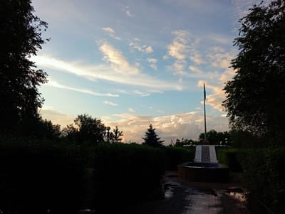 The image depicts an outdoor scene, likely in a park or memorial area, with a flag pole prominently featured in the middle ground. The flag pole is tall and thin, with a flag that appears to be light green and blue, suggesting it may be the flag of Kazakhstan. The flag pole stands on a raised platform accessible by a set of stairs, which are illuminated by lamps on either side.
In the foreground, a circular fountain is visible. The ground surrounding the fountain and leading up to it is paved with bricks, and there are puddles of water on the ground, indicating recent rain or watering. The area is heavily landscaped with dense green bushes and trees, some of which are silhouetted against the sky.
The sky is a mixture of blue and white clouds, with hints of orange and yellow suggesting it is either sunrise or sunset. The lighting conditions indicate it's late afternoon or early evening. There are no people visible in the image.
The location is identified as Qobda, Kazakhstan. The overall atmosphere is serene and perhaps commemorative, given the presence of the flag pole and memorial-like structure.