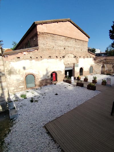 An outdoor scene features an ancient, weathered brick building under a clear, bright blue sky, with a crescent moon faintly visible. The building, likely historical, displays a mix of red brickwork and deteriorating light-colored plaster, revealing its age. It has a gabled roofline and several arched window openings, some fitted with glass. In one prominent arched opening, a red Turkish flag with a white star and crescent hangs vertically. The main entrance, a darker rectangular opening, is located centrally on the building's facade.

In front of the building lies a neatly arranged courtyard area. The ground is predominantly covered with white gravel, interspersed with small patches of green vegetation. Several rectangular wooden planters containing small green plants are symmetrically placed on the gravel, leading towards the building's entrance. A smooth wooden deck or walkway occupies the right side and foreground of the image, contrasting with the light gravel.

Near the entrance, a dark informational sign is visible. Further to the right, another white sign clearly displays universal "no" symbols, indicating restrictions such as no smoking, no food or drinks, and no pets. The scene is illuminated by bright daylight, casting distinct shadows, suggesting either morning or late afternoon. The overall impression is one of a well-maintained historical site, possibly a museum or public area, enjoying a serene, sunny day in Iznik, Turkey. No people are visible.