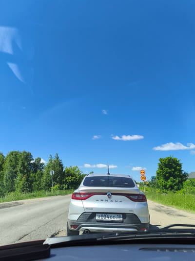 The image is taken from inside a vehicle, looking forward at a silver Renault Arkana car driving on a paved road in the city of Yagul, Russia. The car is in the foreground, slightly to the right of the center. The road is cracked and appears to be in disrepair. To the left and right of the road are lush green trees and vegetation, indicative of a rural or suburban environment. The sky is a clear, bright blue with a few scattered white clouds. There are two circular traffic signs visible on the right side of the road. The upper sign depicts two red circles with horizontal bars, signifying a restriction or prohibition, while the lower sign shows a red circle with the number "70" inside, indicating a speed limit of 70 kilometers per hour. The license plate of the Renault Arkana is partially visible, displaying Russian characters and numbers. The overall scene suggests a sunny day in a scenic, possibly remote, location.