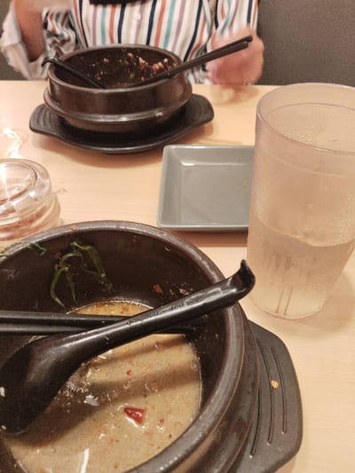 A partially eaten bowl of soup sits on a wooden table, next to an empty bowl, a glass of water, a small gray plate, and a glass container. The soup bowl is dark brown, with a light brown broth inside, some green pieces of vegetation, and a sliver of red pepper. A black spoon is resting on top of the soup. The empty bowl also appears to be dark brown and is slightly tilted to the side. The glass of water is clear, filled with a yellow liquid, and is sitting on a placemat. The gray plate is empty. The glass container is empty. The background appears to be a wood-paneled wall.  The image was taken in the town of Telukjambe, Indonesia. The coordinates are -6.301 latitude and 107.278 longitude.
