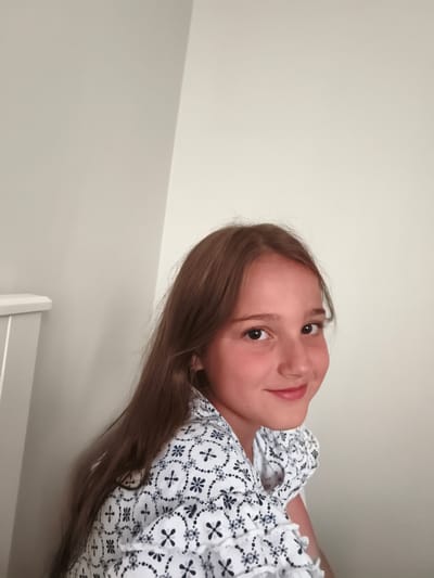 This media file is a medium shot of a young girl with long, brown hair. She is looking directly at the camera and smiling. She is wearing a white shirt with a black floral pattern. The background is a plain, light-colored wall. The lighting suggests it is daytime. There is a white piece of furniture on the left side of the image. The overall mood of the image is cheerful and candid. The girl appears to be in a domestic setting, possibly her home in Dobele, Latvia.