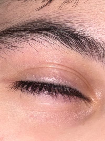 A high-resolution, extreme close-up depicts a human eye, currently closed. The eyelid skin is fair-toned with visible fine texture and subtle folds above the lash line. Dark, thick eyelashes extend from the eyelid, appearing slightly glossy along their length. A dense, dark eyebrow arches above the eyelid, with individual hairs clearly discernible. The immediate surrounding area consists solely of the subject's facial skin, providing no external environmental context. This scene is located in the city of Saint-Étienne, France.