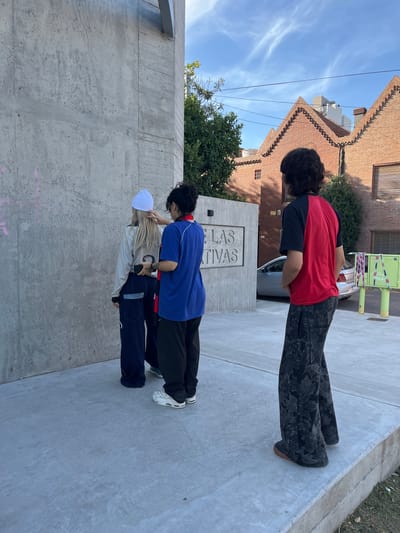 In Bahía Blanca, Argentina, three individuals stand on a concrete sidewalk in front of a concrete wall bearing the inscription "LAS TIVAS." The first person is wearing a red and black shirt with dark patterned pants. The second person is wearing a blue shirt, black pants, and white sneakers. The third person wears a white hat, a cream-colored sweater, and dark pants with white accents. They are all facing towards the inscription on the wall. A building is visible behind the first person, and a car is parked nearby.