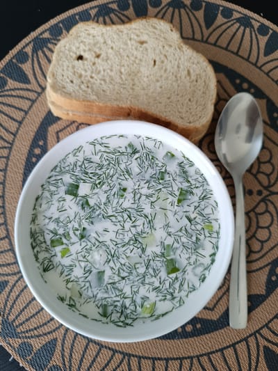 A white bowl of cold soup, likely okroshka, filled with finely chopped dill and cucumber, is centered on a brown placemat with a circular, patterned design. To the left of the bowl are two slices of white bread. To the right of the bowl is a silver spoon. The soup has a creamy white base, with the green dill and cucumber creating a speckled appearance. The bread is lightly toasted. The scene appears to be a casual meal, possibly lunch, indoors. There is no visible text. The overall impression is one of a refreshing and simple meal.