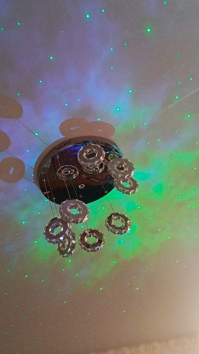 A modern chandelier with crystal rings hangs from a ceiling. The ceiling is illuminated by a starry projector, casting green and purple light patterns. Shadows of the chandelier's crystal rings are visible on the ceiling. The image is an indoor shot, likely in a residential setting in Zvenigorod, Russia.